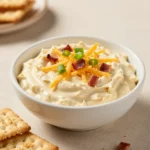 A bowl of creamy Crack Dip topped with shredded cheese and chopped green onions, served with crackers and fresh vegetables.